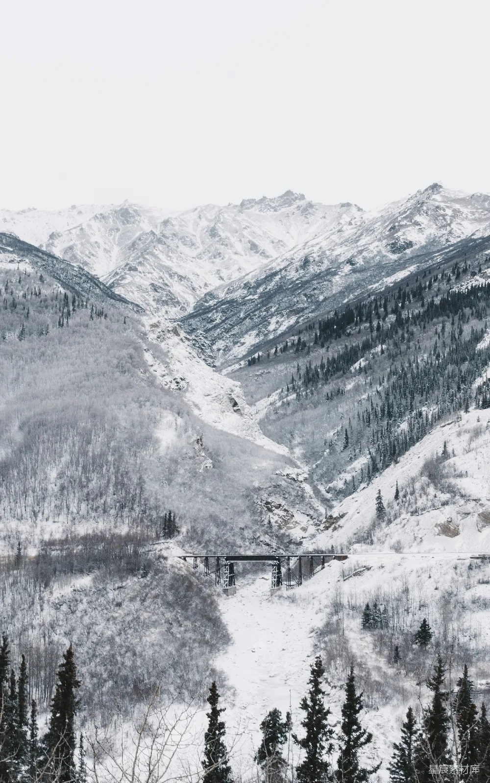 高清摄影 黑桥周围是被白雪覆盖的山峦，在白茫茫的天空下。