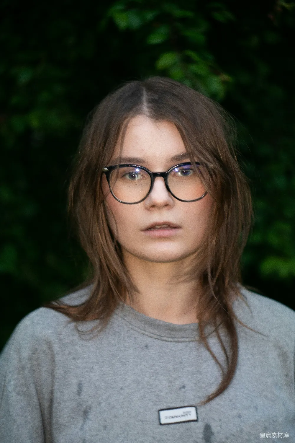 高清摄影 戴着眼镜、棕色头发的年轻女子站在绿色植被前。