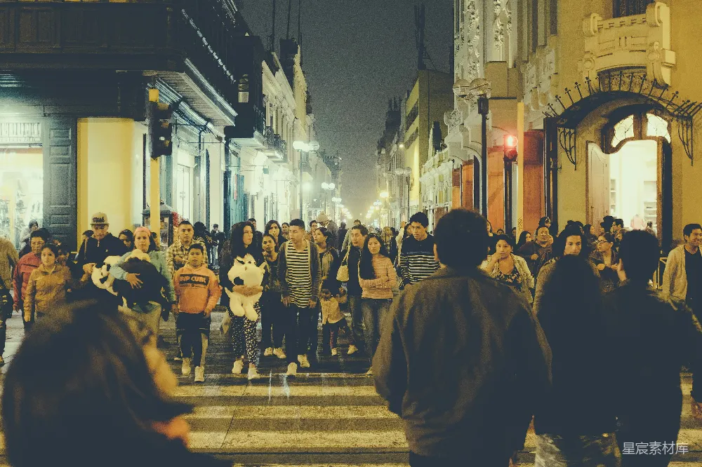 图片展示了一个夜晚城市中的繁忙十字路口，街道两旁是装饰华丽的建筑，灯光昏黄而温暖，人们在人行道上忙碌地行走，有的抱着孩子，有的背着包，呈现出一幅充满活力的都市夜景。