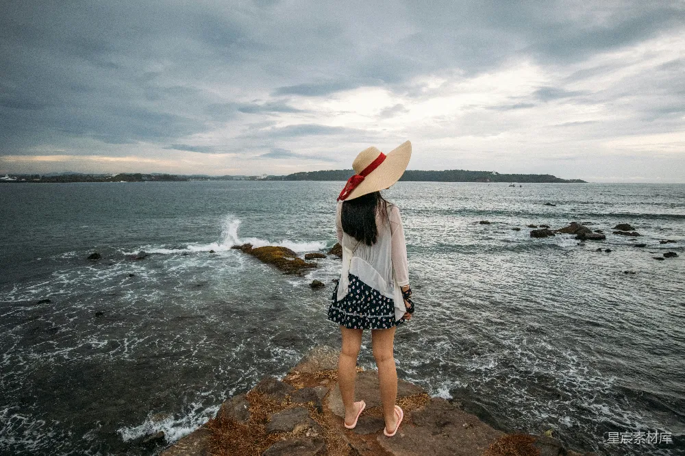 高清摄影  戴着帽子的女人在岩石海岸观看着海浪。