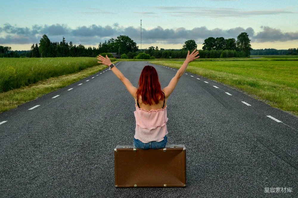 高清摄影  女人举起双臂坐在行李箱旁，处于开阔的道路上。