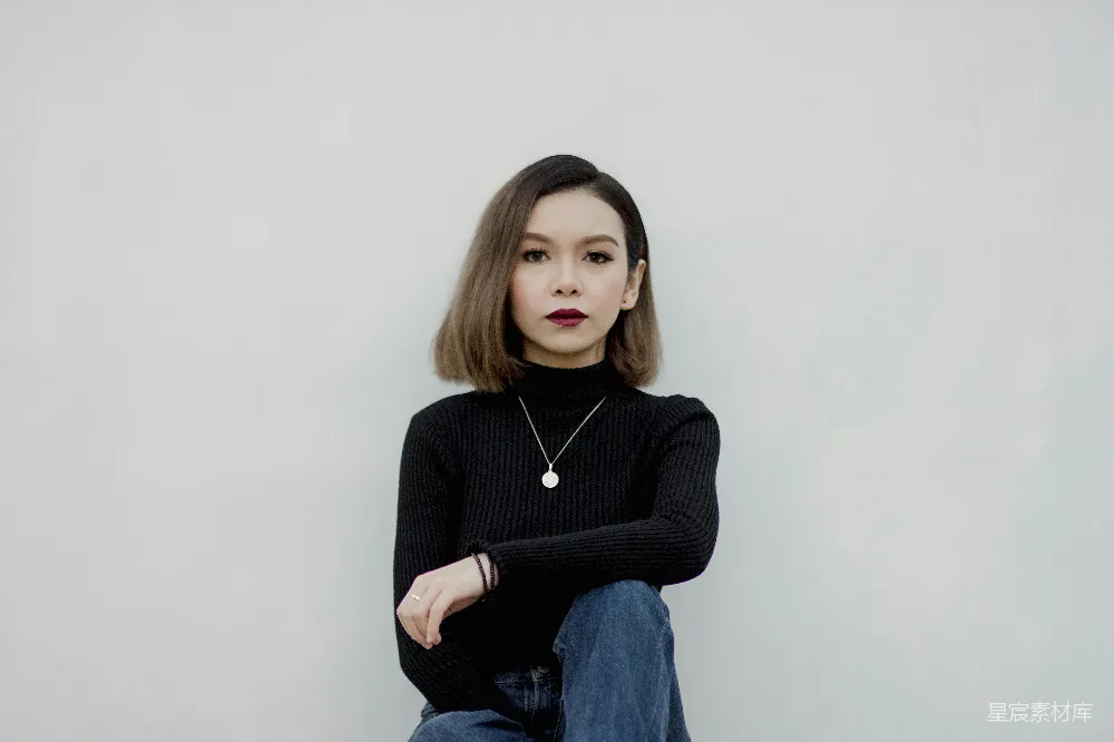 这张图片展示了一位穿着简约黑色高领毛衣的女性，搭配简洁的蓝色牛仔裤，背景为纯白色，营造出一种干净、简约的视觉效果。她微微侧身，双手交叉在背后，呈现出一种自信且随性的姿态，适合用于个人写真、时尚杂志或简约风格的广告素材。