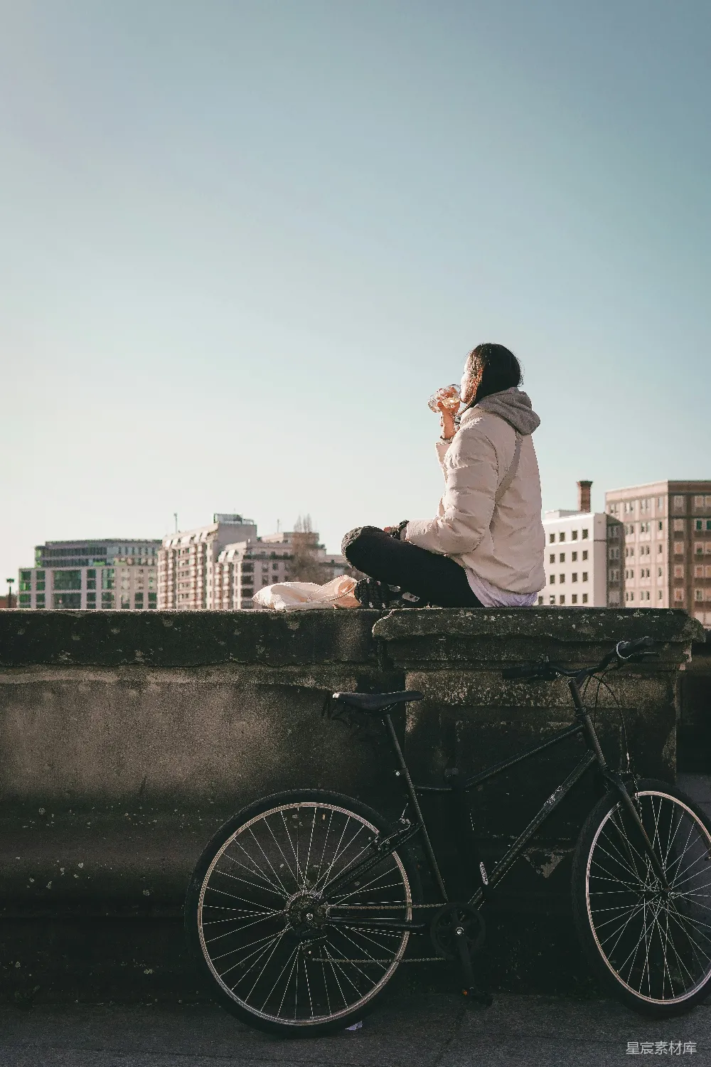 高清摄影 女人坐在墙角，脚踏自行车，背景是城市。