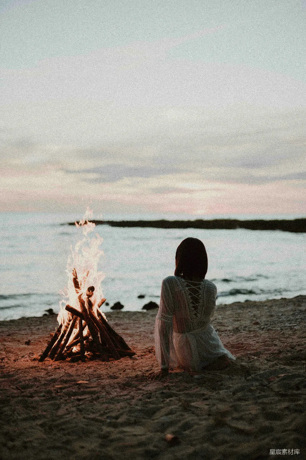 高清摄影  一个女人坐在海滩前的篝火旁。