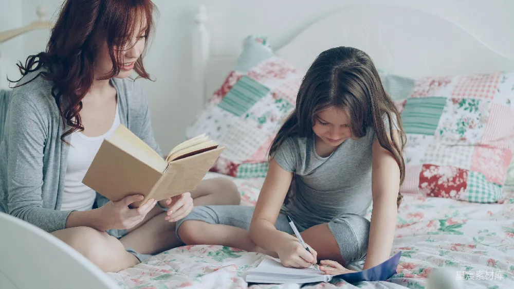 细心的母亲帮助她的小可爱女儿完成小学作业。慈爱的妈妈在书上阅读，女孩则在笔记本上做笔记，她们一起坐在家里的温馨卧室里。