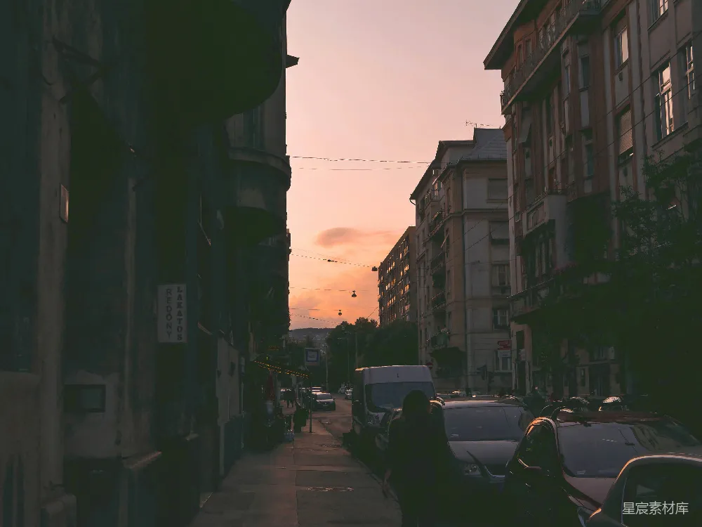 白天，在橙色天空下，人们在近建筑物的小道上行走，道路两旁有各种车辆。