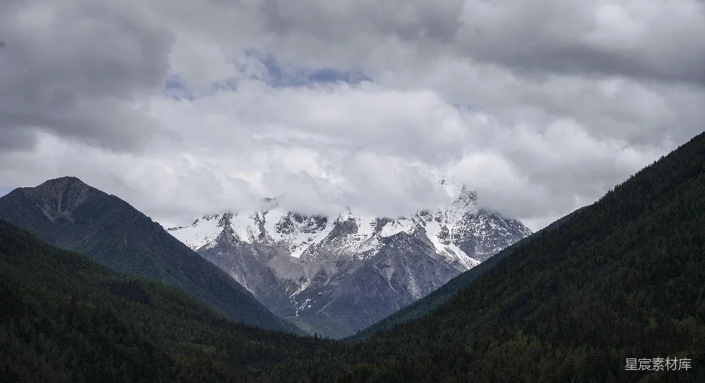 高清摄影  白天阴云密布的天空下，山峦被白雪覆盖，近旁绿树成荫。