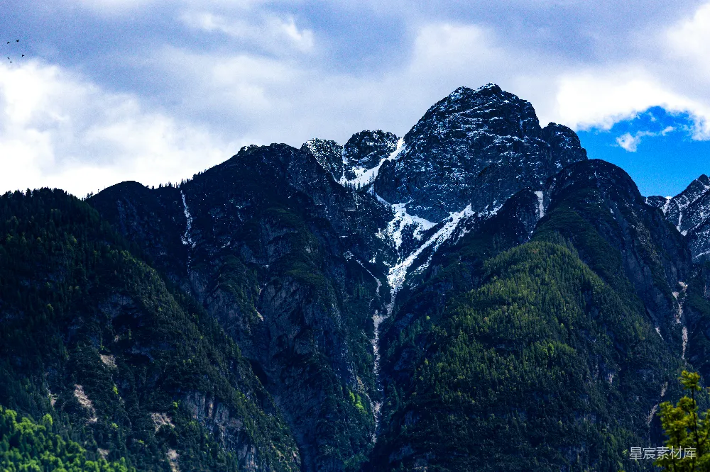 高清摄影  云层密布的天空下，白雪覆盖的山峰旁有绿树环绕。