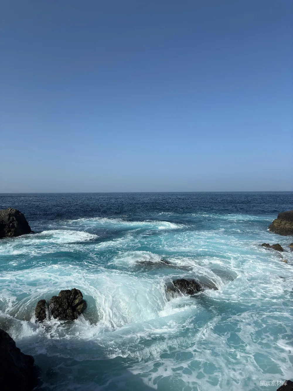 高清摄影  波浪在清澈的蓝天下拍打着岩石。