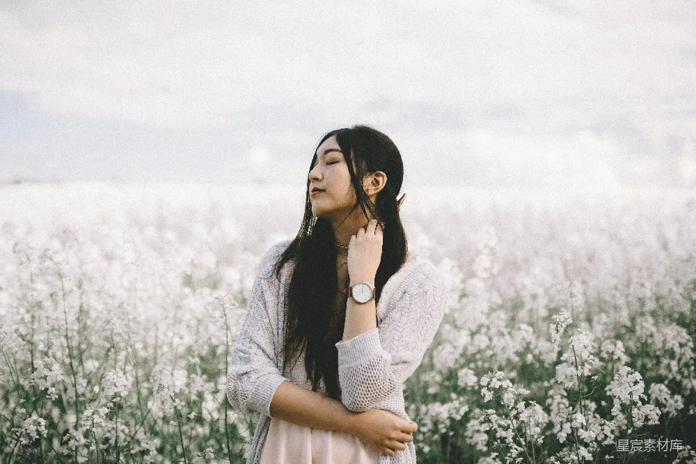 高清摄影  一名穿着桃色内搭和灰色针织开衫的女子站在白天的白色花丛后面拍摄。