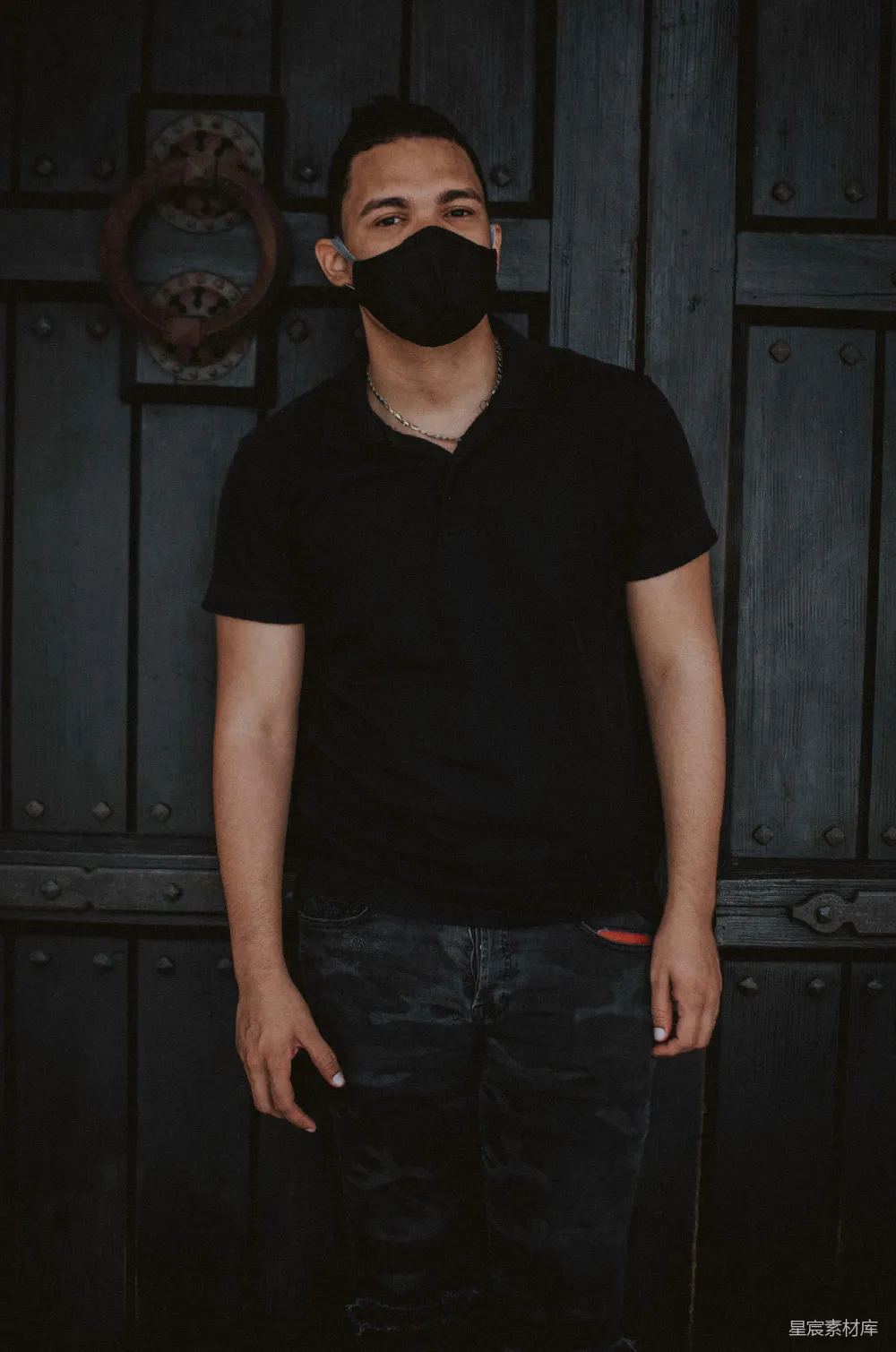 高清摄影  穿着黑色V领T恤和蓝色牛仔裤的男人站在黑色木门前。