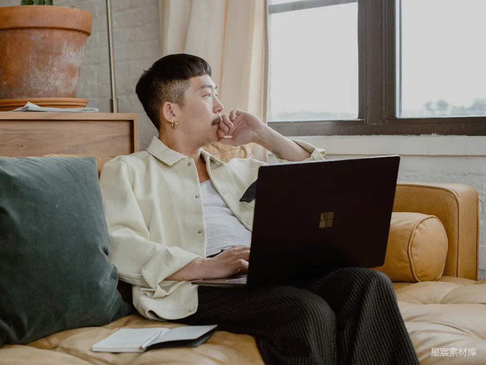 高清摄影  穿着白色衬衫和黑色裤子的男人坐在沙发上，使用Surface笔记本电脑。