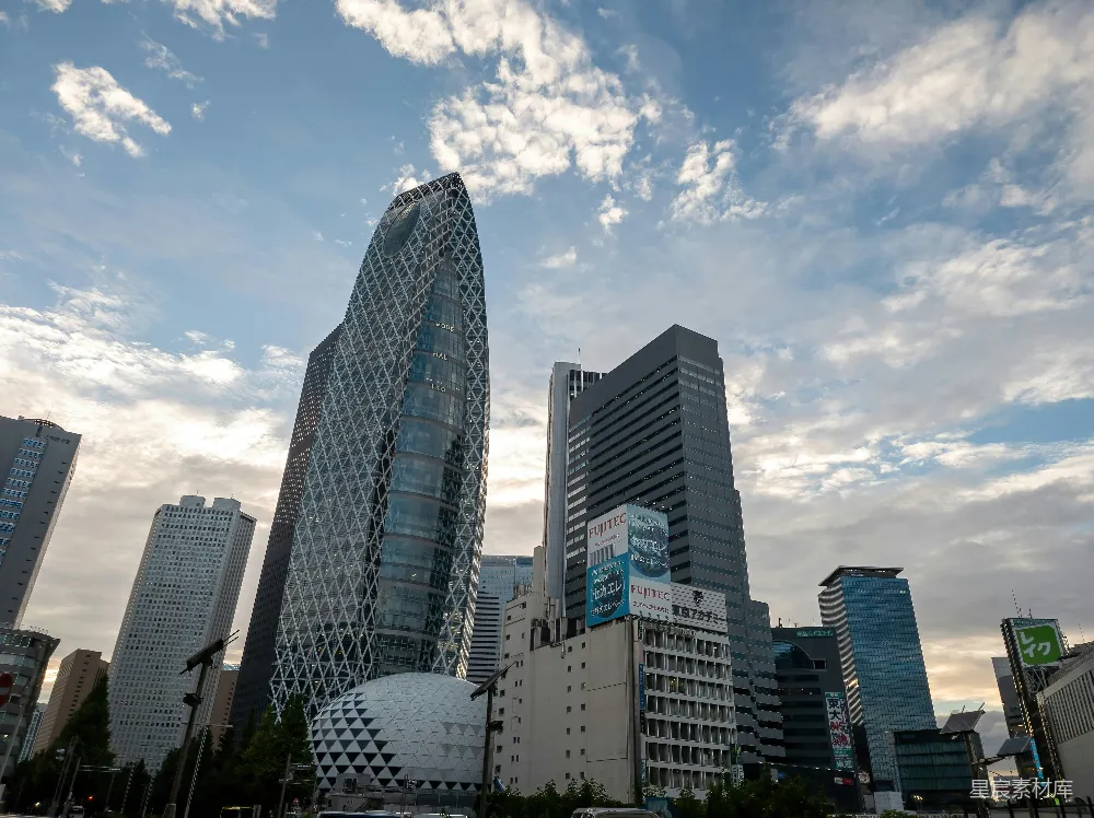 这张图片展示了东京的标志性建筑之一，独特的几何图案摩天大楼在蓝天白云的映衬下显得格外醒目。周围的建筑风格各异，展现了东京作为国际大都市的多元化建筑风貌。适合用于商业、旅游或城市规划相关的内容。