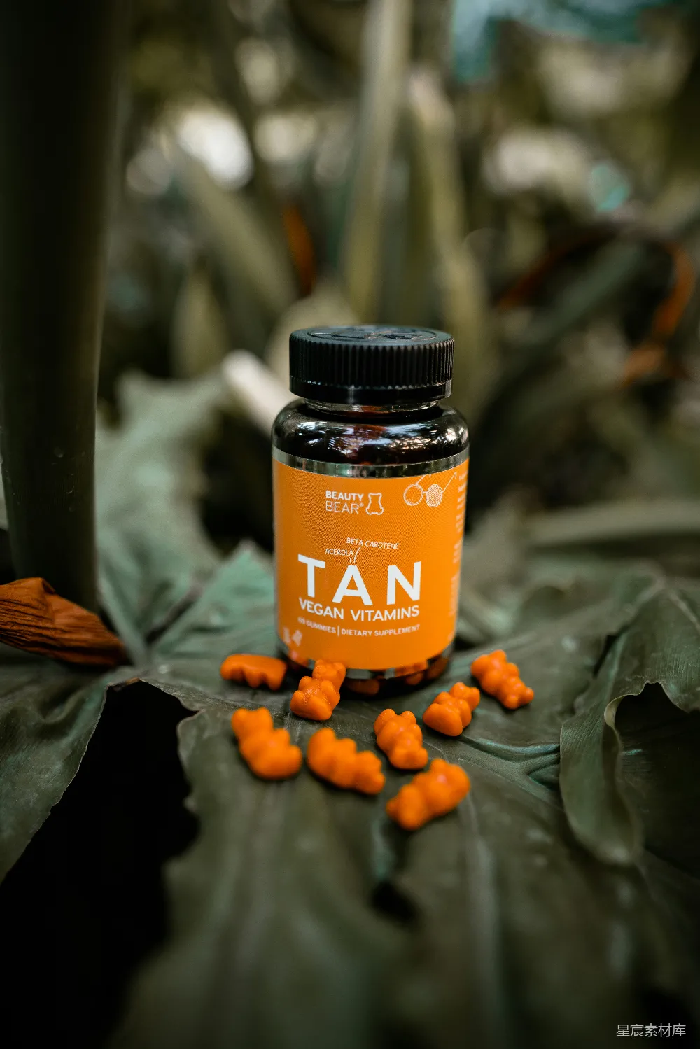 图片展示了一瓶标有“TAN”字样的有机素食维生素补剂，瓶身橙色，瓶盖黑色，瓶内有橙色的维生素软糖。背景是模糊的绿色植物，突显出产品自然、健康的特质。适合用于日常健康保健，特别是注重素食和健康生活方式的人群。