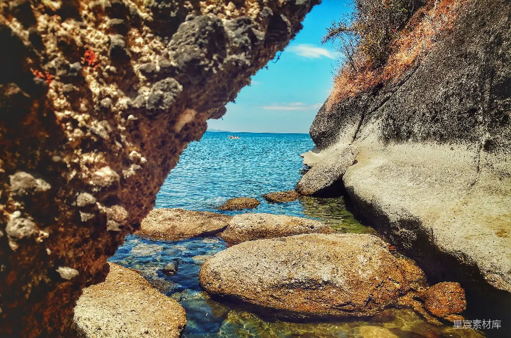 这张图片展示了岩石缝隙中的一片海景。清澈的海水在阳光下闪耀，岩石表面粗糙，形成了天然的框架，将海景尽收眼底。背景是广阔的蓝天和远处的山丘，整个画面充满了宁静与自然的美。适合用作旅游或自然风光的图片素材。
