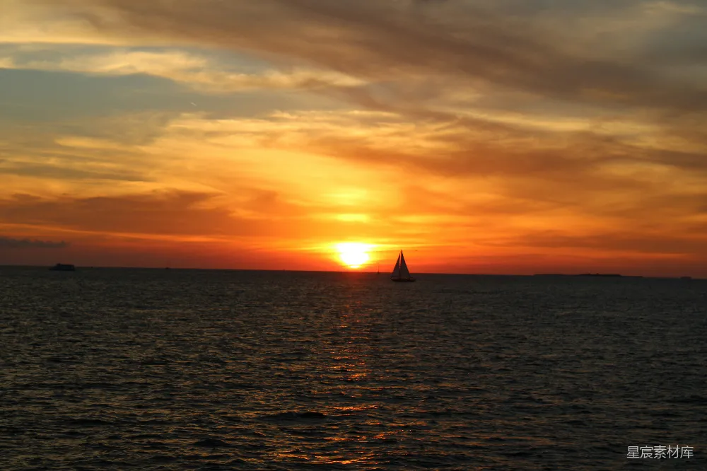 这张图片展示了一片宁静的海景，夕阳即将落下，天空被染成了金黄色和橙红色。海面上波光粼粼，一艘帆船在海面上静谧地航行，给人一种宁静而美好的感觉。适合用作旅行、自然风光或日落主题的素材。
