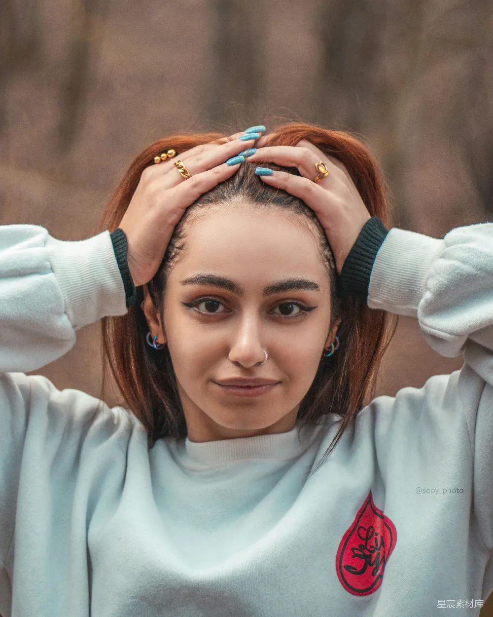 这张图片展示了一位年轻女性，她双手握着自己的头，显得非常自信和有个性。她穿着一件带有红色徽章的浅蓝色卫衣，背景模糊，突出了人物的主体。整体风格充满青春活力，适合用于展示年轻一代的时尚和个性。