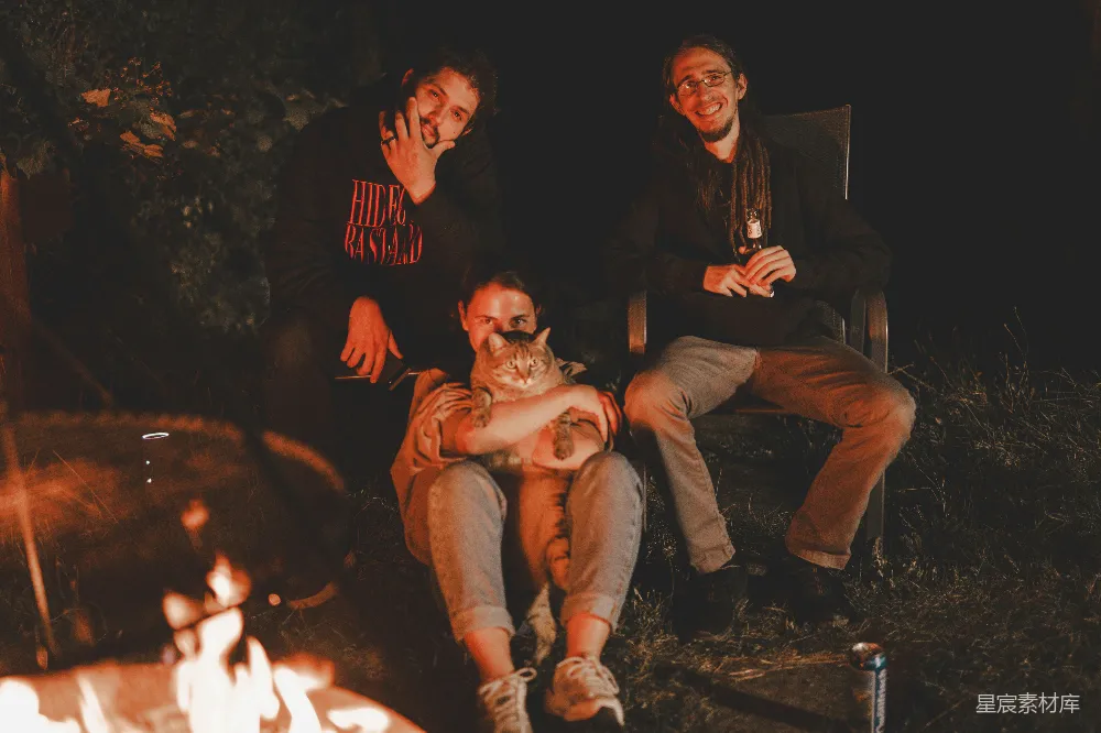 三人在夜晚的篝火旁聚会，其中一人抱着一只猫，氛围温馨而亲密。篝火的温暖光芒照亮了他们，背景中可以看到灌木丛，营造出一种自然而放松的氛围。这张图片适合用作朋友间的温馨时刻、户外活动的分享或是宠物爱好者的展示。