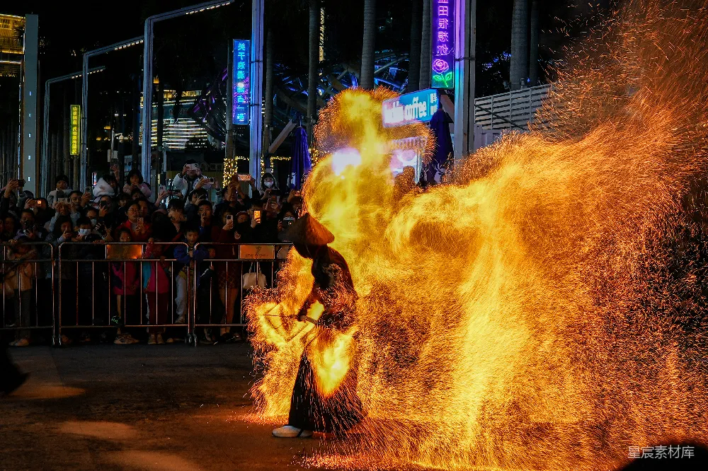 夜晚的街头，一位表演者在灯光下舞动着火光斑斓的狮子，吸引着无数观众驻足观看。火光与观众的手机灯光交织在一起，形成一幅生动的夜市景象。
