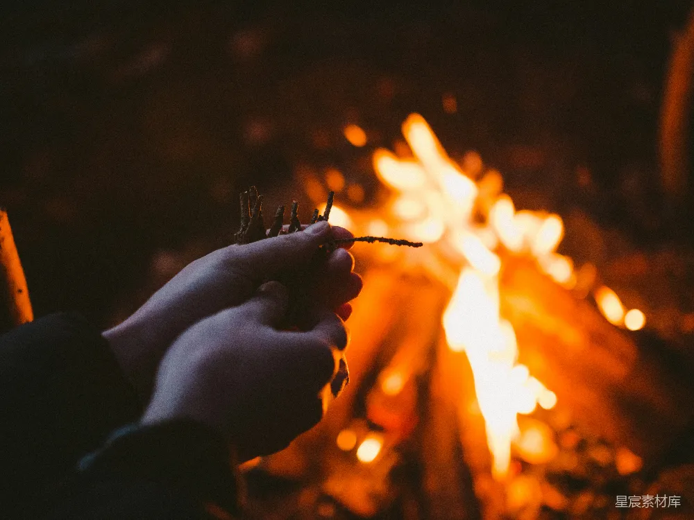 一只手拿着干柴靠近熊熊燃烧的篝火，背景是模糊的火光，营造出一种温馨而宁静的氛围。这幅图片适合用于表达户外露营、篝火晚会等场景，强调人与自然的亲近和生活的简单美好。