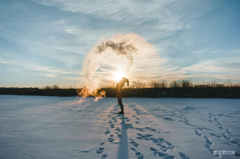 图片中，一个穿着冬季服装的人站在雪地上，阳光透过他呼出的寒气，形成一个美丽的圆形光环。背景是落日的余晖和远处的树木，地面上清晰地留下了脚印。整个场景展现了冬日寒冷但又充满诗意的美丽。