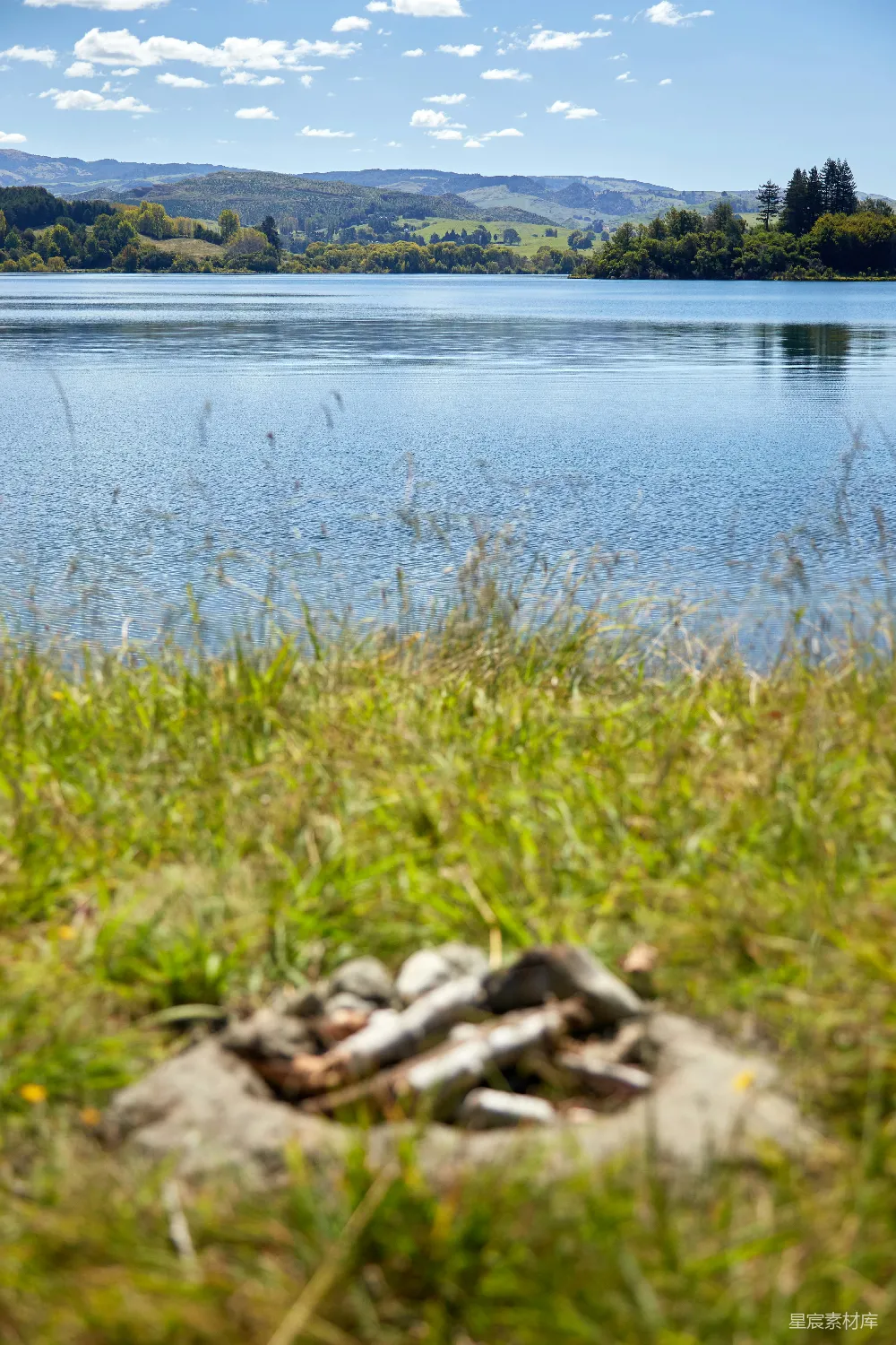 这张图片展示了宁静的湖泊景色，前景是绿色的草地和一些木头，背景则是广阔的湖泊和远处连绵的山脉。天空中散布着几朵白云，阳光明媚，整个画面显得非常宁静和自然。适合用于描述户外休闲、自然摄影或旅行目的地。