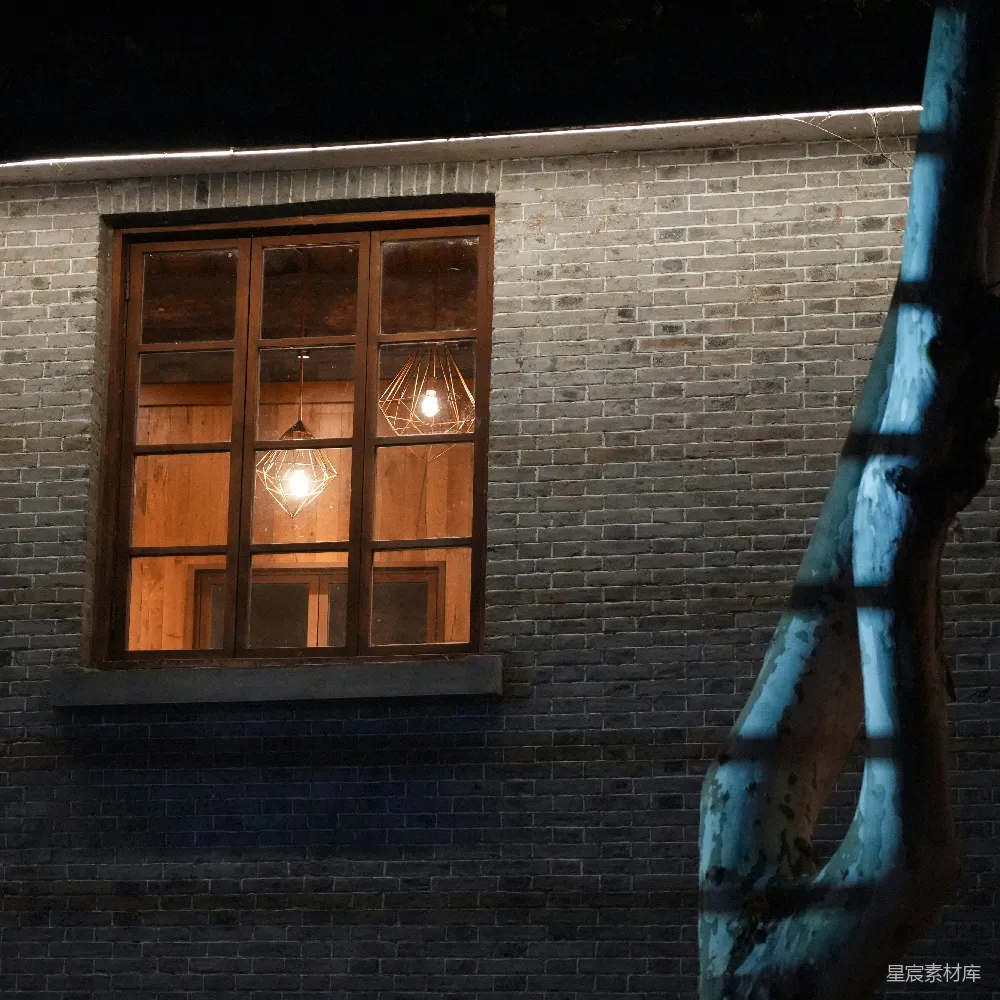 夜晚的砖墙窗户内透出温暖的灯光，窗外挂着一个独特的几何灯饰，砖墙表面略显斑驳，一旁的蓝色雕塑为画面增添了艺术感。这幅图像展现了城市夜晚的独特氛围，适合用作城市夜景、复古风格或艺术摄影的背景素材。