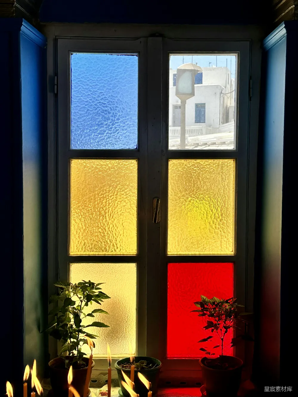 这是一幅展示在室内拍摄的照片，画面中有一扇彩色玻璃的窗户，透过窗户可以看到外面的街道和一盏路灯。窗户下方有几株绿植，其中一盆植物前点燃了几根蜡烛，散发出温暖的光芒。窗户的玻璃呈现出蓝色、黄色和红色，形成了一种独特的视觉效果，给人一种宁静而美