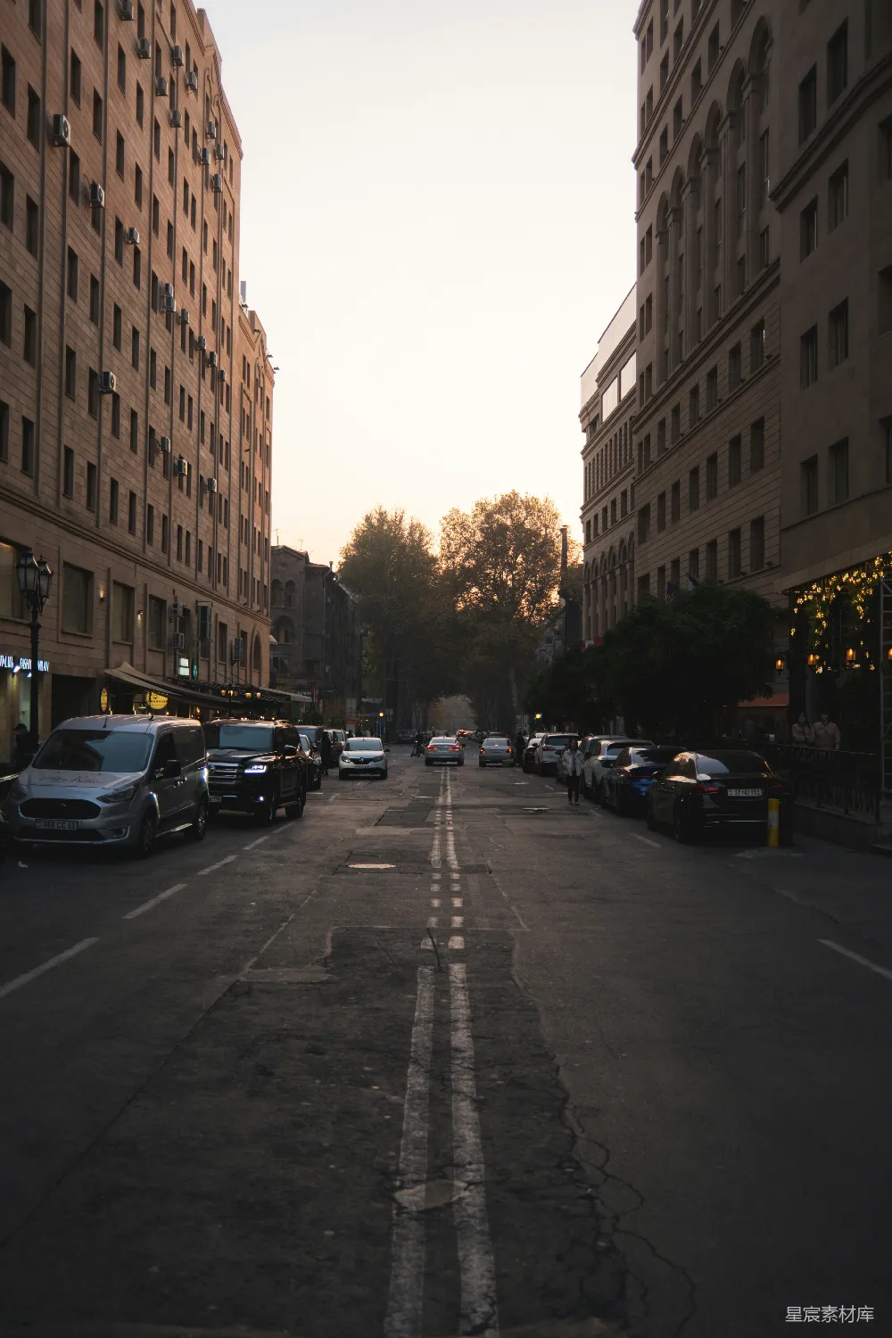 这张图片展示了一条城市中的街道，黄昏时分的光线柔和地洒在路面上，树木和建筑物在背景中显得宁静而和谐。街道两旁停满了车辆，远处的灯光开始点亮，营造出一种平和而略带孤寂的氛围。适合用于描述城市生活中的一个安静片段，或是用于与城市黄昏相关的视觉素