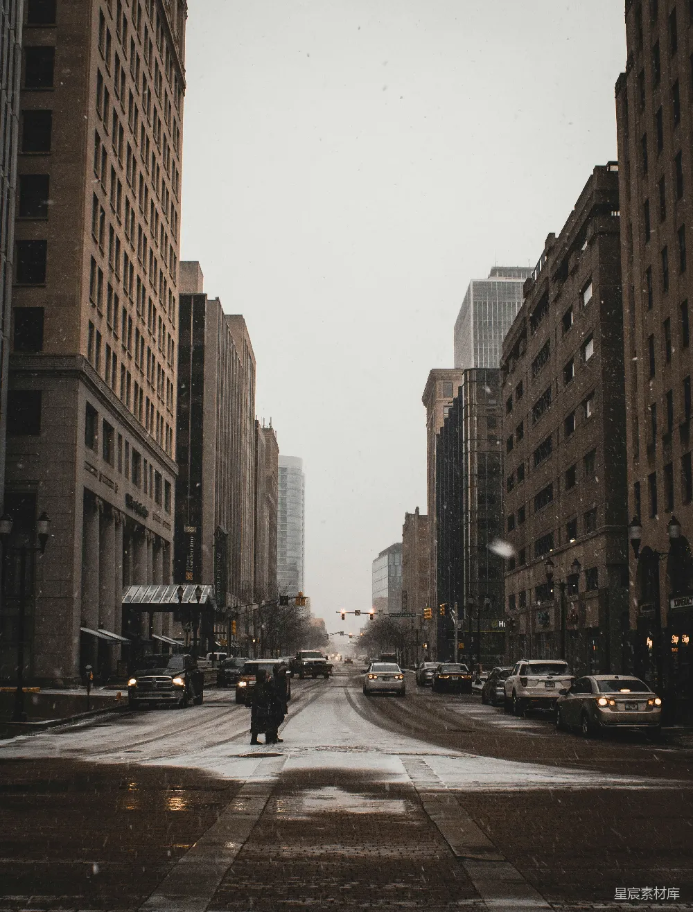 图片展示了一座城市的街道，在冬日的雪景中显得格外宁静。两侧是高大的建筑物，街道上车辆稀疏，行人稀少。雪花缓缓飘落，给整个场景增添了一层淡淡的诗意。整体色调偏冷，给人一种安静而神秘的感觉。