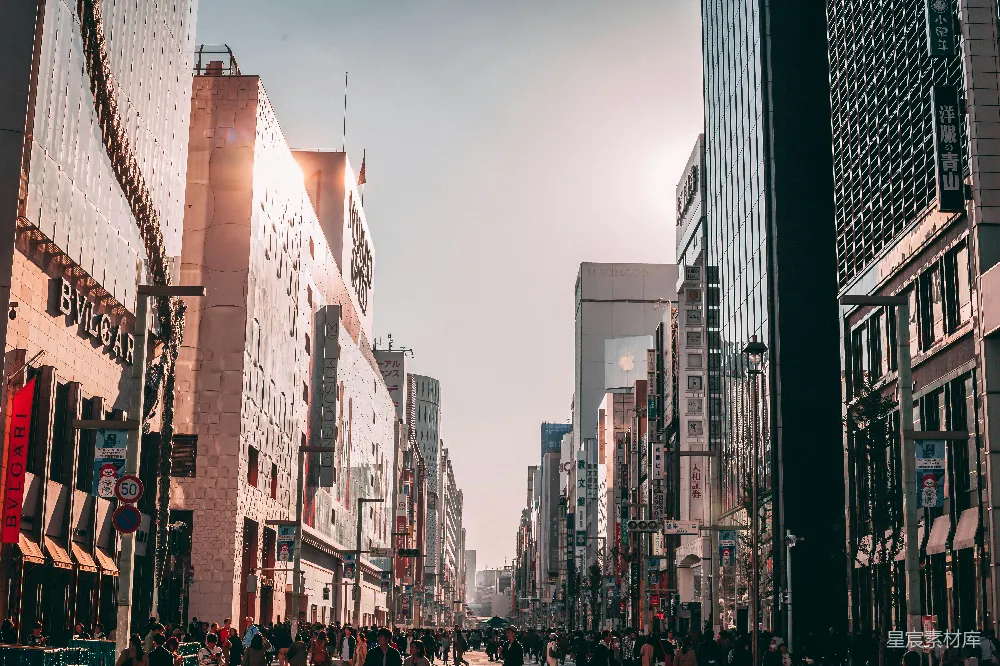 这张图片展示了一条繁忙的城市街道，两旁是高大的建筑，街道上人来人往，显得非常热闹。阳光透过建筑物投射下来，营造出一种温暖的氛围。街道两旁的招牌和广告牌清晰可见，显示出这是一个商业区。适合用于城市生活、商业或旅游相关的内容展示。