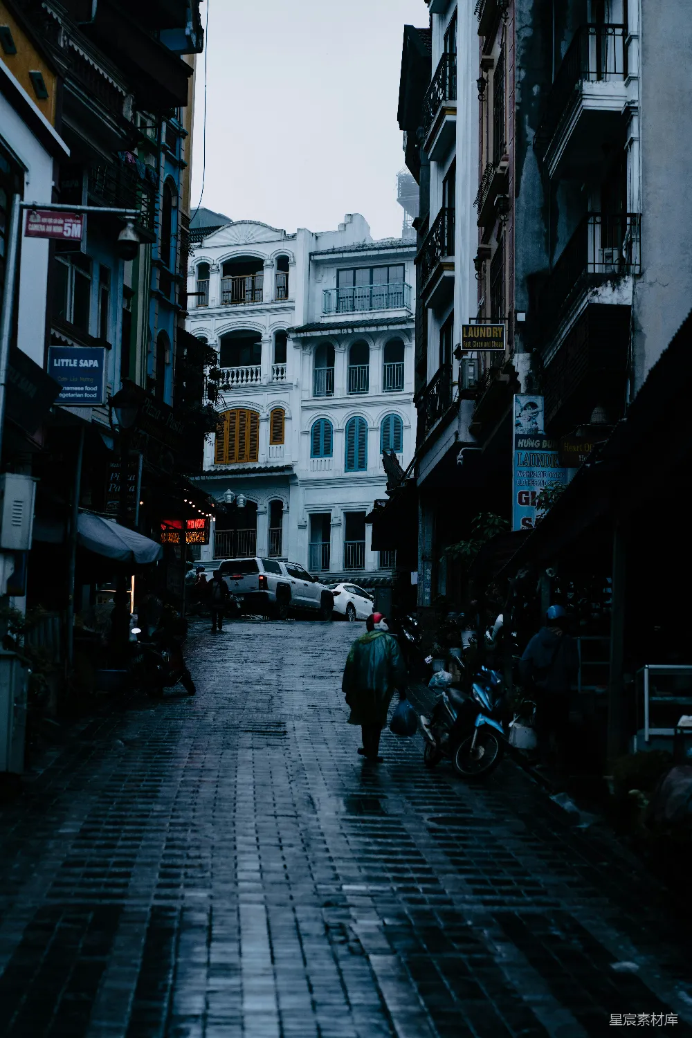 图片展示了一条铺满石砖的老街，两侧是带有阳台的建筑，街道上有一位穿着雨衣的人在行走。天空阴沉，街道上有些积水，远处停着几辆汽车。整体色调偏冷，给人一种宁静而略带忧郁的感觉。适合用于描述城市生活、怀旧或雨天场景的素材。