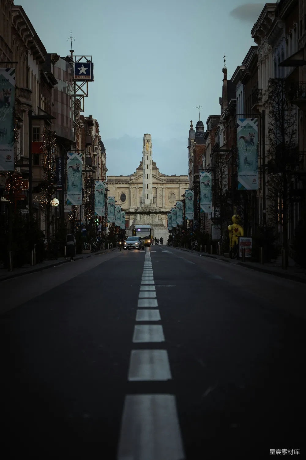 这张图片展示了一条宁静的欧洲城市街道，街道两旁是装饰着节日彩灯的建筑，中间的纪念碑在黄昏时分显得格外庄重。道路上的车辆稀少，给人一种安静祥和的感觉。适合用于城市风光、旅行摄影或城市生活主题。