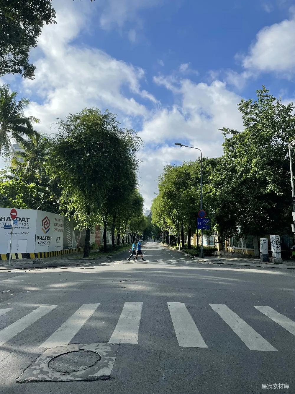 这张图片展示了一条城市街道，两旁是茂密的绿树，阳光洒在街道上，形成斑驳的光影。街道上只有两位行人，天空湛蓝，云朵稀疏，给人一种宁静而舒适的感觉。适合用作城市生活、自然美景或休闲时光的背景图。