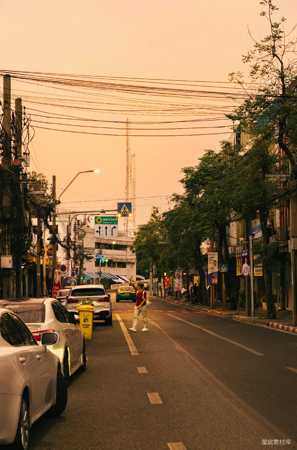 这张图片展示了一条城市中的街道，夕阳的余晖洒在街道上，给整个场景增添了一层温暖的色调。街道两旁是停放的车辆和行走的行人，背景中可以看到一些商店和树木，远处的天线上方还有一座高塔。整个画面给人一种宁静而舒适的感觉，仿佛是在一个悠闲的傍晚时分，