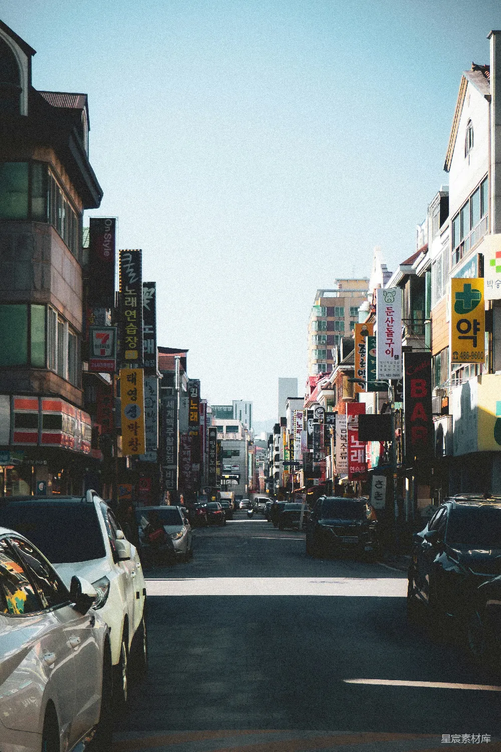 这张图片展示了一条充满活力的韩国商业街。两侧是各种商店和广告牌，街道上停着几辆车，天空晴朗，阳光明媚。街道两旁的建筑风格多样，显示出浓厚的地域特色。适合用于展示城市生活、旅游或商业主题的内容。