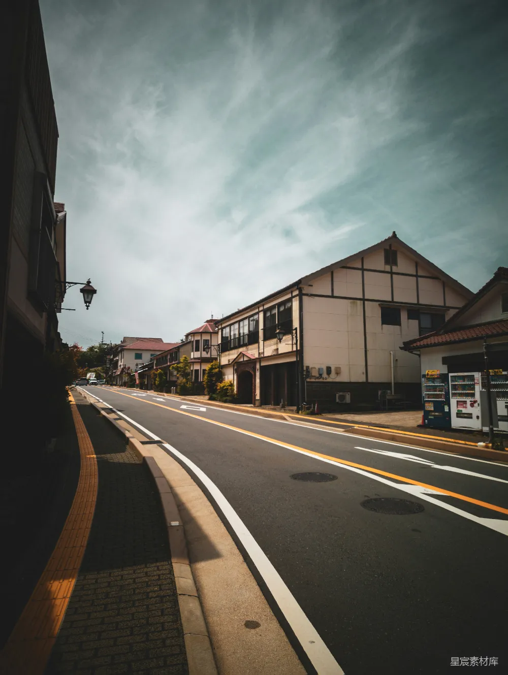 这张图片展示了一条宁静的小镇街道，两旁是典型的日式建筑，天空中飘着几朵白云。街道干净整洁，路边有一个自动贩卖机，整体给人一种平和而悠闲的感觉。适合用于展示日本小镇的日常生活场景。