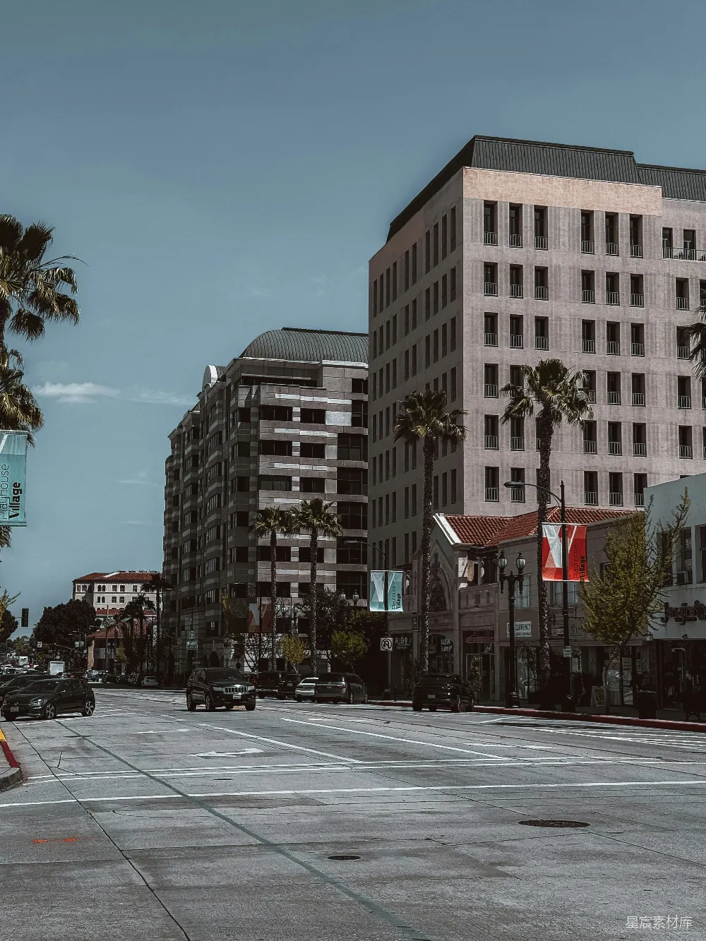 这张图片展示了一个阳光明媚的城市街景，前景是一条宽阔的街道，道路上有几辆汽车在行驶。街道两旁是现代风格的建筑，其中一栋建筑顶部有独特的圆形设计，另一栋建筑则显得更为传统。街道上种满了高大的棕榈树，为城市增添了一份热带风情。天空晴朗，蓝天与建
