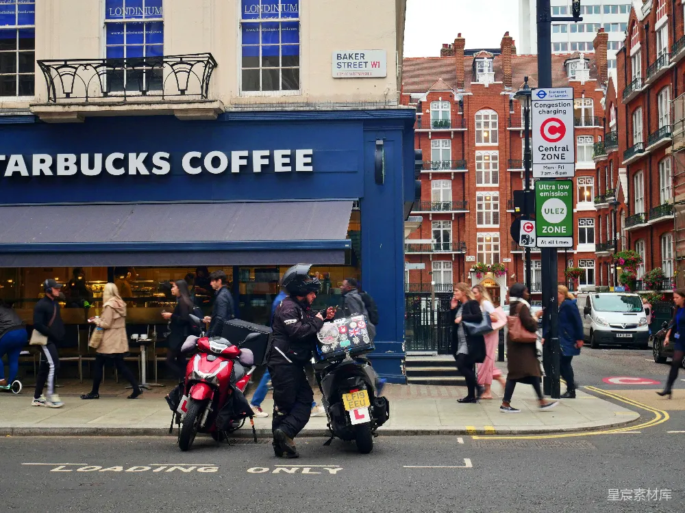图片展示了伦敦贝克街的一角，前景是一家星巴克咖啡店，背景是红砖建筑和行人。画面中可以看到一个外卖员骑着摩托车穿过街道，周围还有行人和路标，营造出一个充满活力的都市氛围。