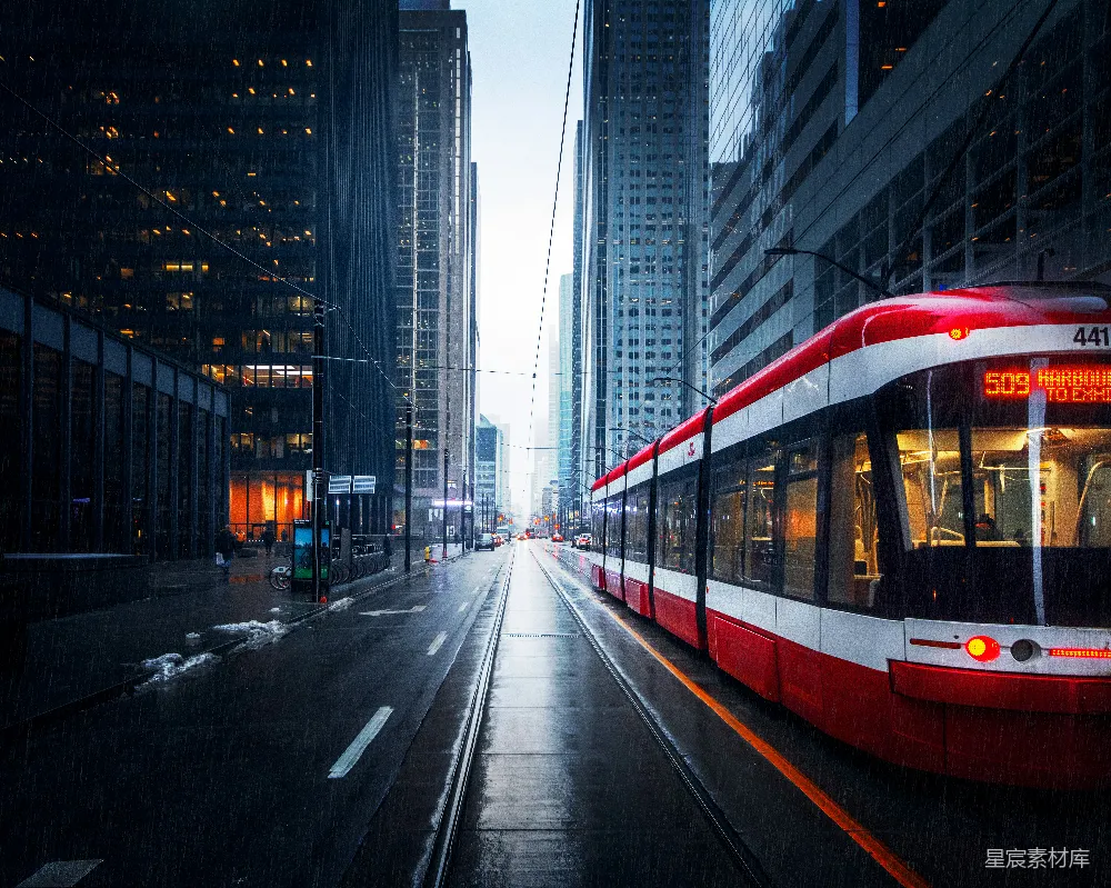 这张图片展示了一辆红色的现代电车在雨中行驶的城市街景。街道两旁是高楼大厦，反射着雨滴，营造出一种湿润而宁静的氛围。电车的红色在灰暗的雨中显得格外醒目，车窗映出车内乘客的身影，增添了生活气息。场景适合用于现代城市生活、交通出行或雨景主题的背景