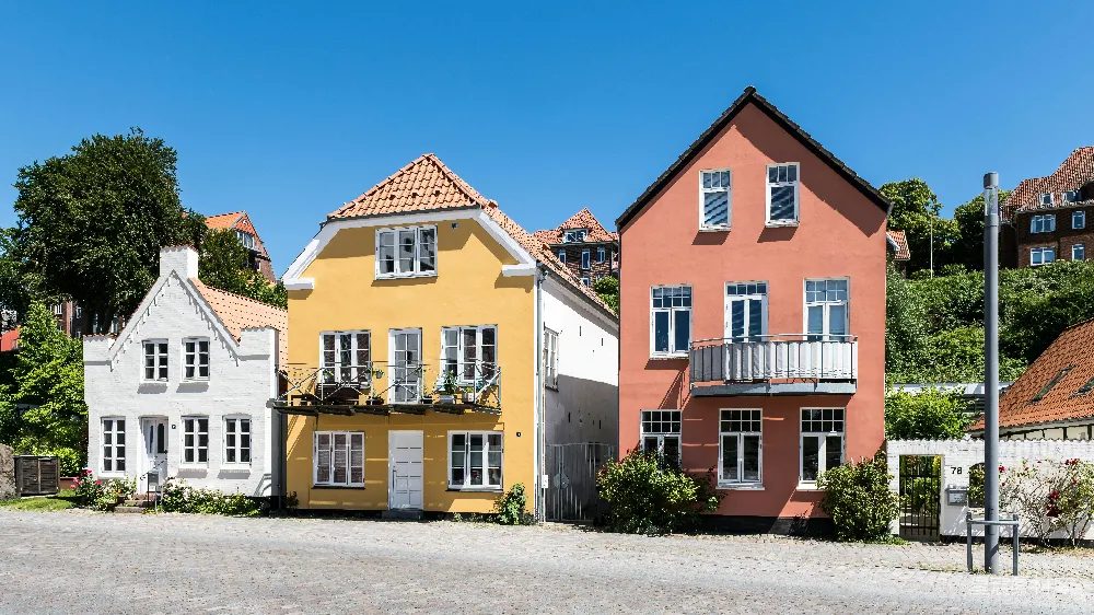 图片展示了一排色彩斑斓的丹麦住宅，黄色、白色和粉红色的房屋在蓝天的映衬下显得格外明亮。房屋设计简洁，窗户排列整齐，周围种植了一些绿色植物，营造出宁静和谐的氛围。适合用于旅行、建筑和家居设计相关的内容展示。