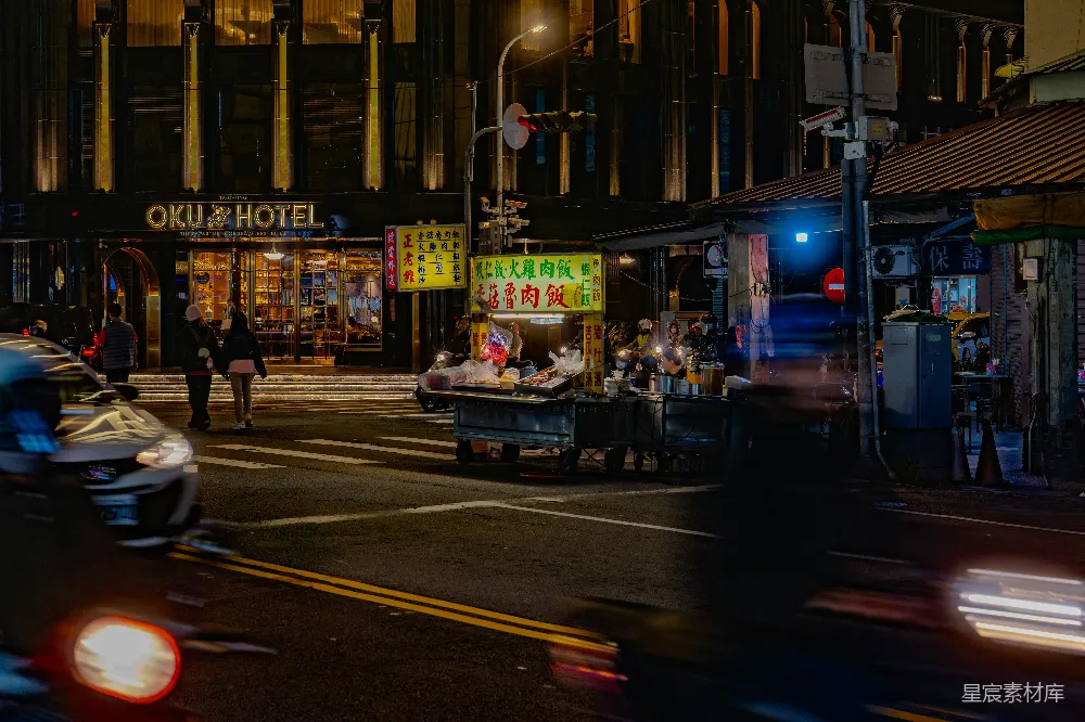 这张图片展示了夜晚的城市街景，画面中有一个摆放在路边的小吃摊，摊位上的招牌亮着黄色的灯光，吸引着路人的目光。背景中，一家名为“OKU HOTEL”的酒店在灯光的映衬下显得格外醒目。街道上车辆的尾灯在夜色中形成一道道光带，增添了画面的动感。整