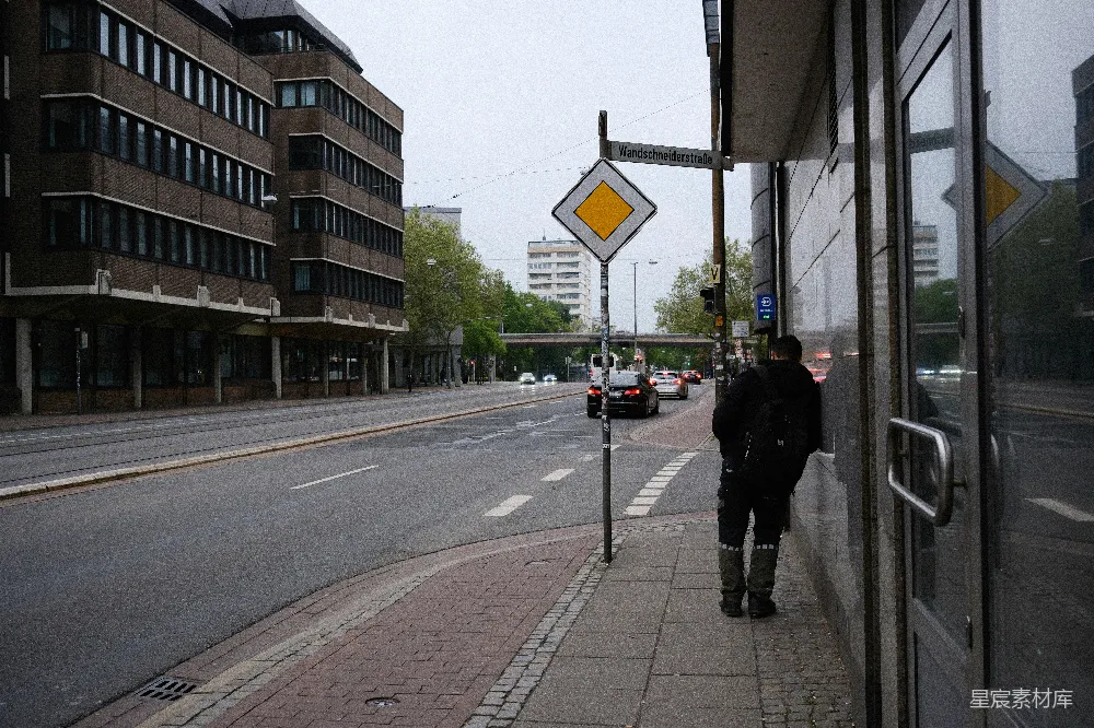 这张图片展示了一条城市的街道，画面中有一个行人在路边等待，街道两旁是现代的建筑。天空阴沉，街道上车流稀少，给人一种宁静的感觉。背景中可以看到一些绿树和远处的建筑物，整体色调偏冷，适合表现城市中的一片宁静时光。
