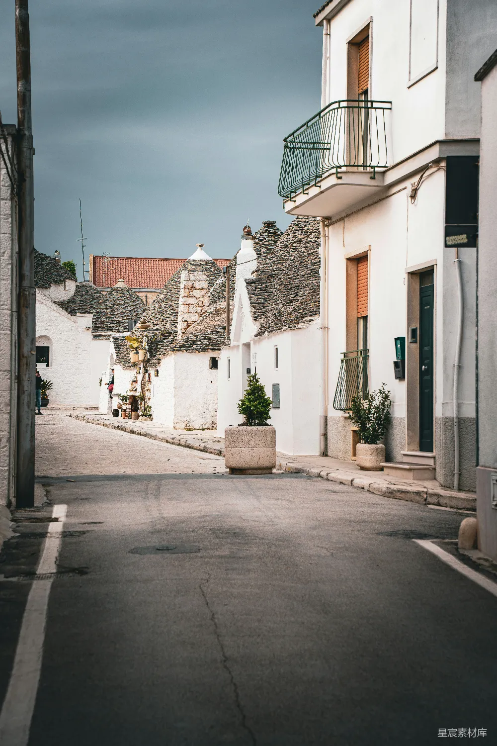 这张图片展示了意大利阿尔贝罗贝洛（Alberobello）的鹅卵石街道，两旁是传统的石屋，屋顶形状独特，给人以宁静和怀旧的感觉。街道上人烟稀少，显得非常安静。