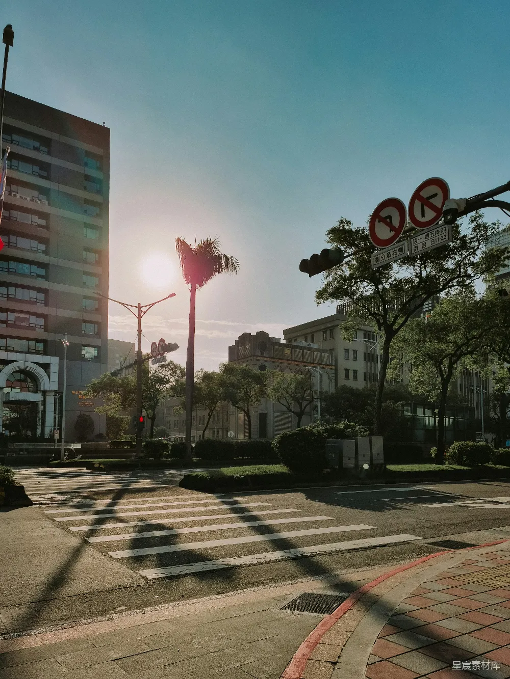 这张图片展示了一个城市的街景，夕阳的余晖洒在街道上，形成美丽的光影效果。画面中有现代化的建筑和传统的建筑物，前景是斑马线和交通标志，背景是高大的树木和蓝天。整个场景给人一种宁静而美好的感觉。