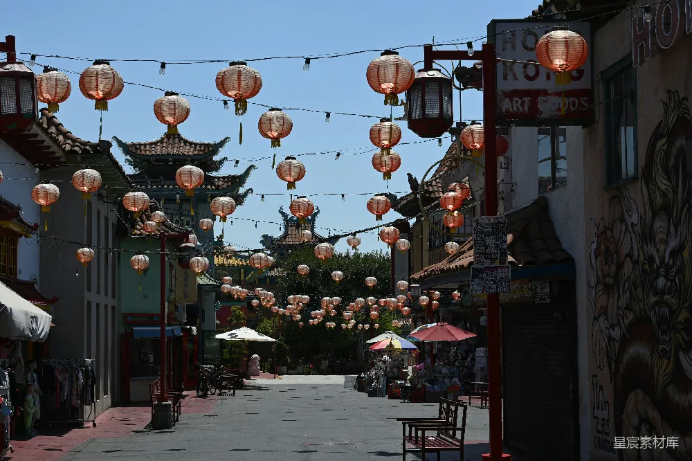 一条街道两旁挂满了红色的灯笼，营造出浓厚的节日氛围。街道左侧有摊贩的帐篷，右侧有店铺和装饰，背景是传统建筑风格的建筑。天空晴朗，阳光明媚，给人一种宁静祥和的感觉。适合拍摄城市文化、节庆场景或休闲旅游的图片。