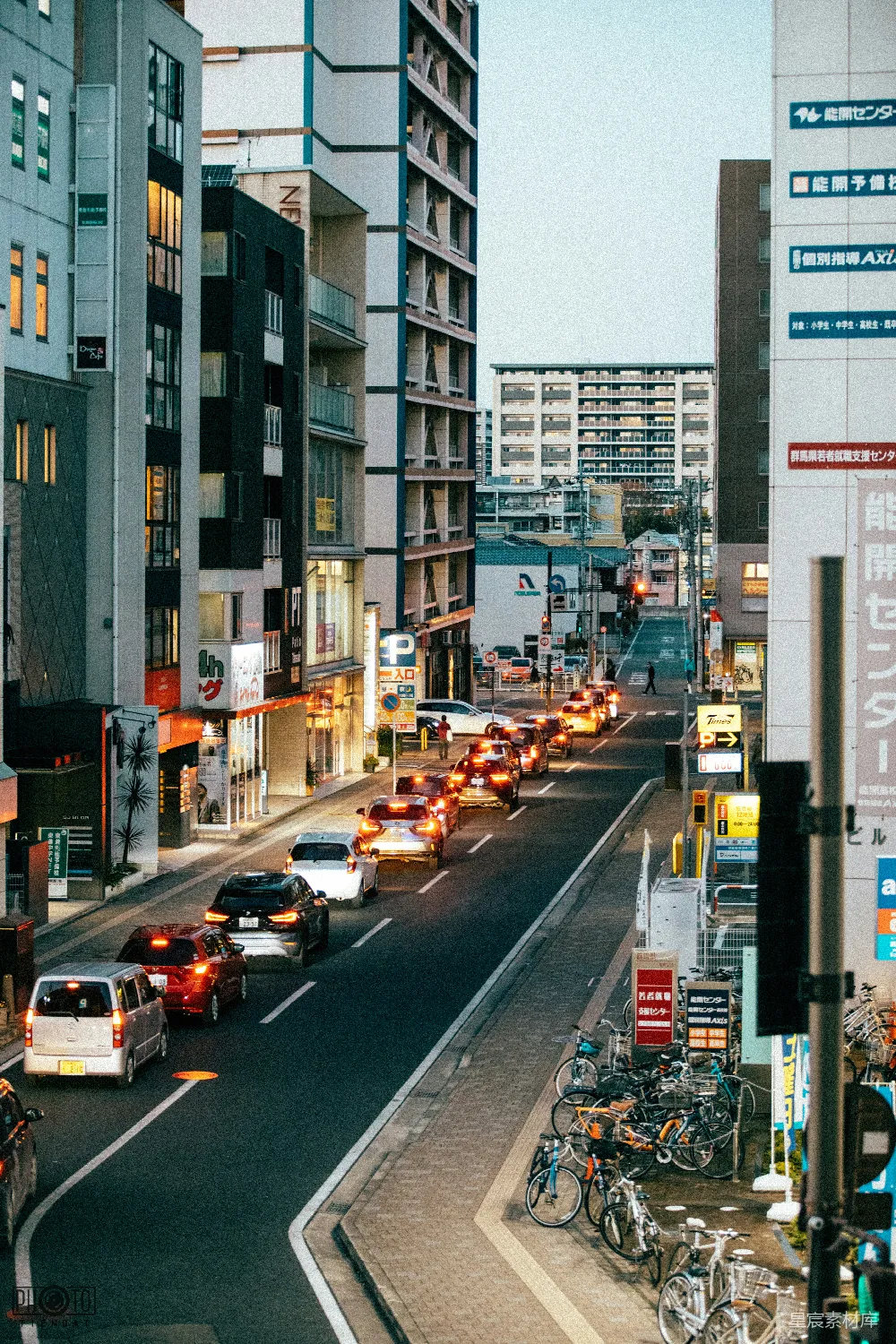 这张图片展示了东京街头的黄昏景色。街道两旁是高大的建筑，街道上车辆川流不息，车灯和路灯的光芒交织在一起，营造出一种宁静而充满活力的氛围。街道一侧停放着许多自行车，显示出东京市民环保出行的普遍习惯。黄昏时分的光线柔和，给整个场景增添了一种温暖