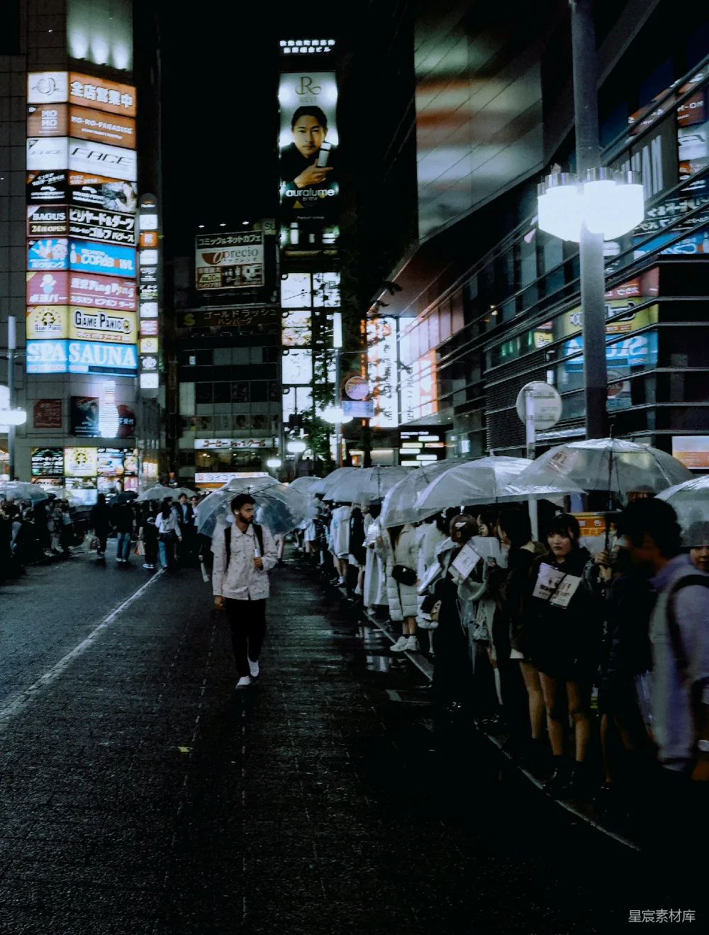 这张图片展示了一个城市的夜景，霓虹灯和广告牌在雨夜中显得格外醒目。街道上，行人撑着透明的雨伞，形成了一道独特的风景线。整个场景充满了现代都市的氛围，同时也带有一种雨夜特有的宁静与神秘。