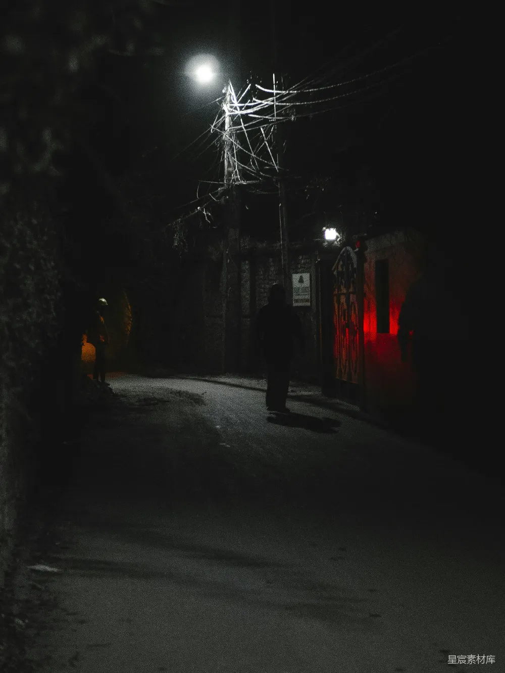 一张夜晚街道的照片，灯光昏暗，街道两旁是电线杆和树木，一个人影孤独地走在街道上，远处的红色灯光为场景增添了一丝神秘感。整体画面给人一种静谧而略带忧郁的感觉。