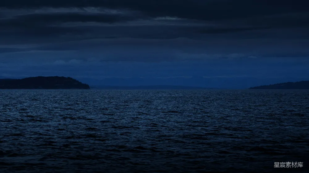 这张图片展示了在夜幕下的宁静海景。天空中布满了深蓝色的云层，海面波光粼粼，远处可以看到陆地的轮廓。整体色调深沉，给人一种宁静而神秘的感觉，适合用作自然风光或夜间场景的背景素材。